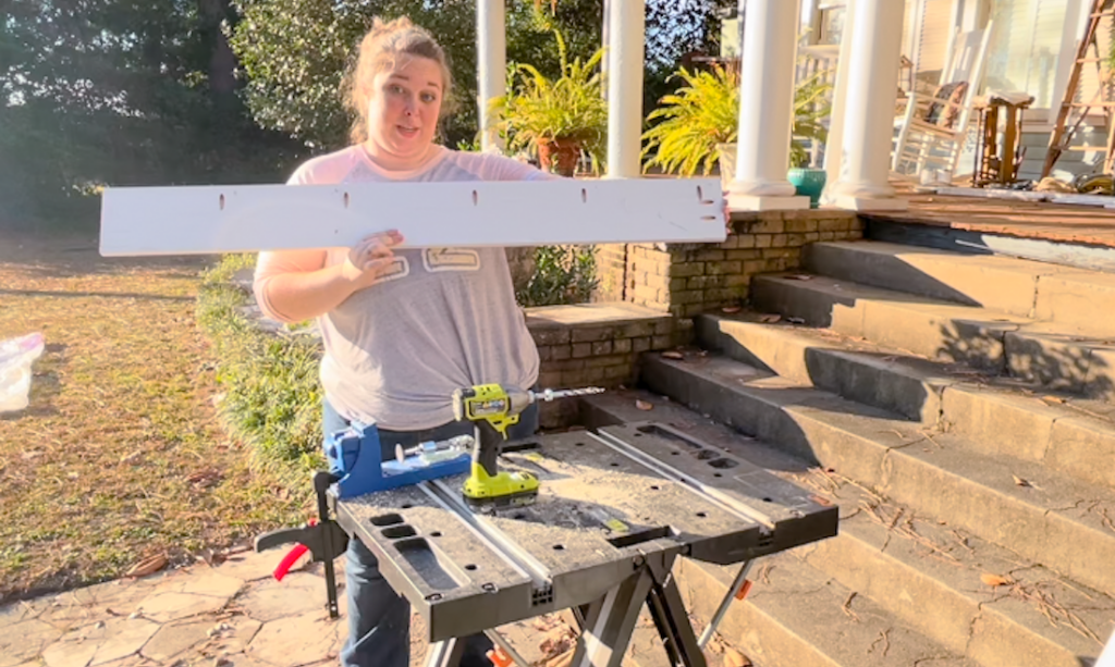 person holding piece of wood up looking at the camera, they are at a work bench